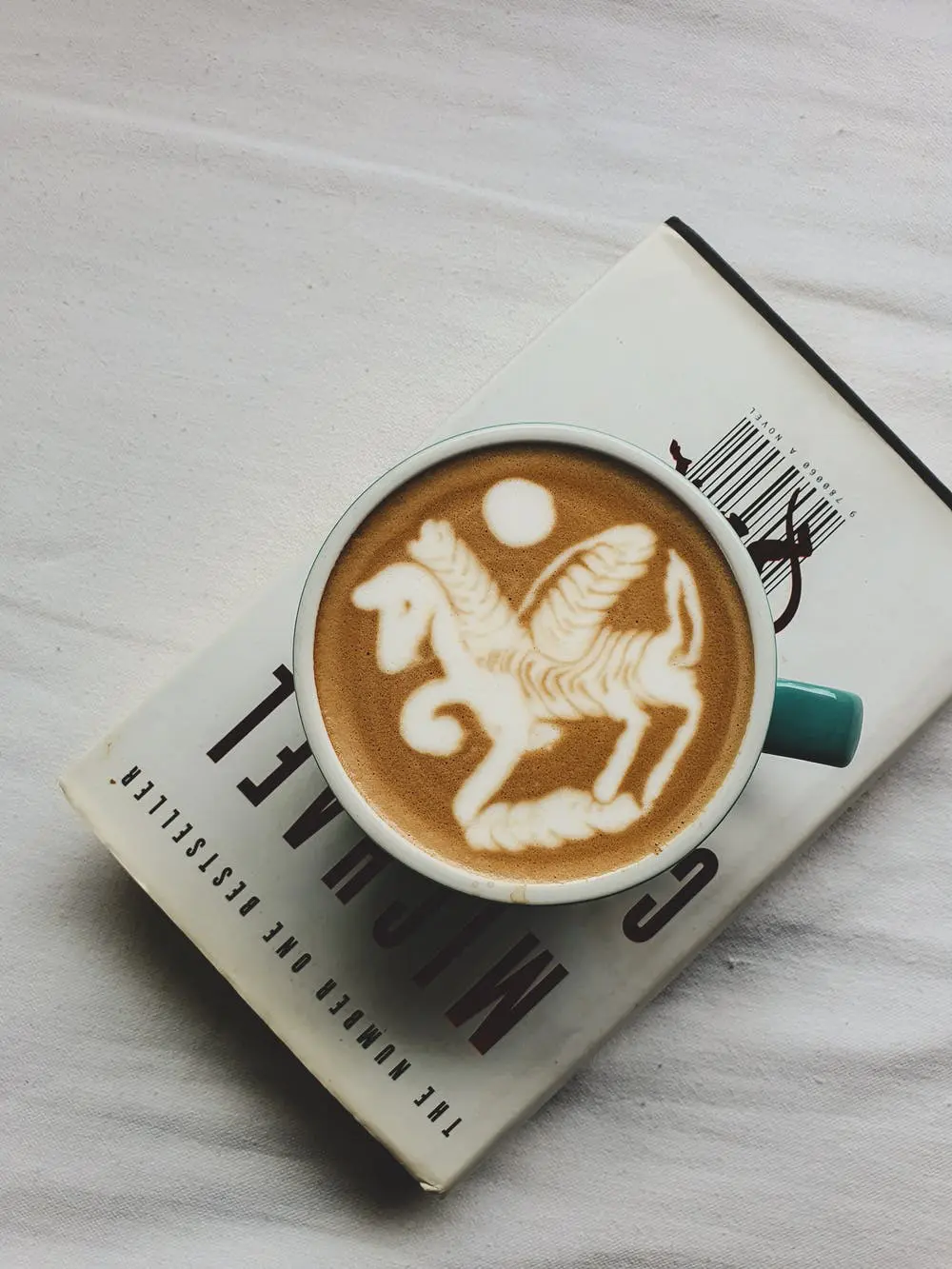 Cup of latte with a white horse-shaped latte art in a turquoise mug resting on a book on a white surface.