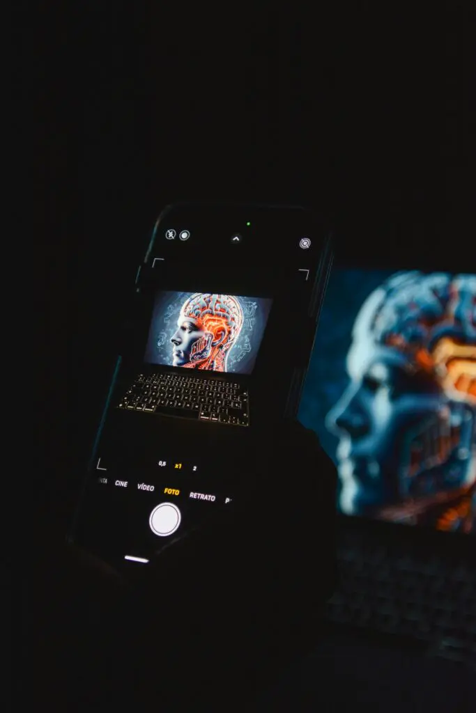 Smartphone screen showing a brain illustration inside a camera app, with a computer monitor displaying a similar image in the background (dark scene).
