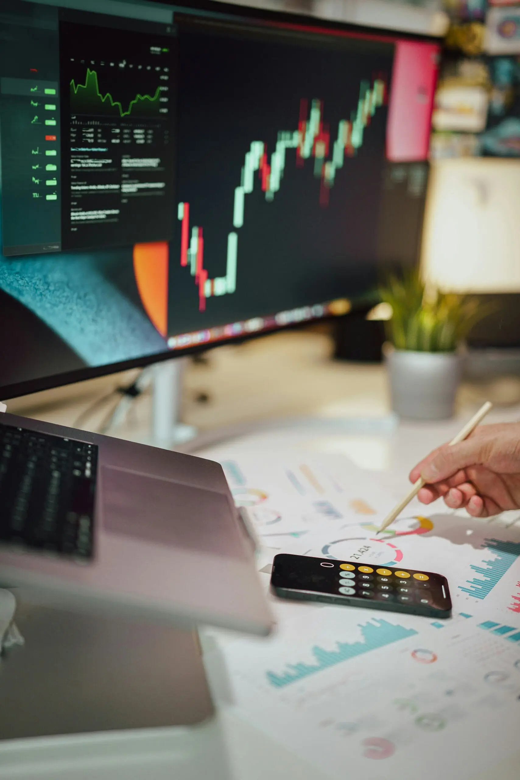 Desk workspace with a monitor showing stock charts, a hand annotating printed charts with a pencil, and a calculator nearby.