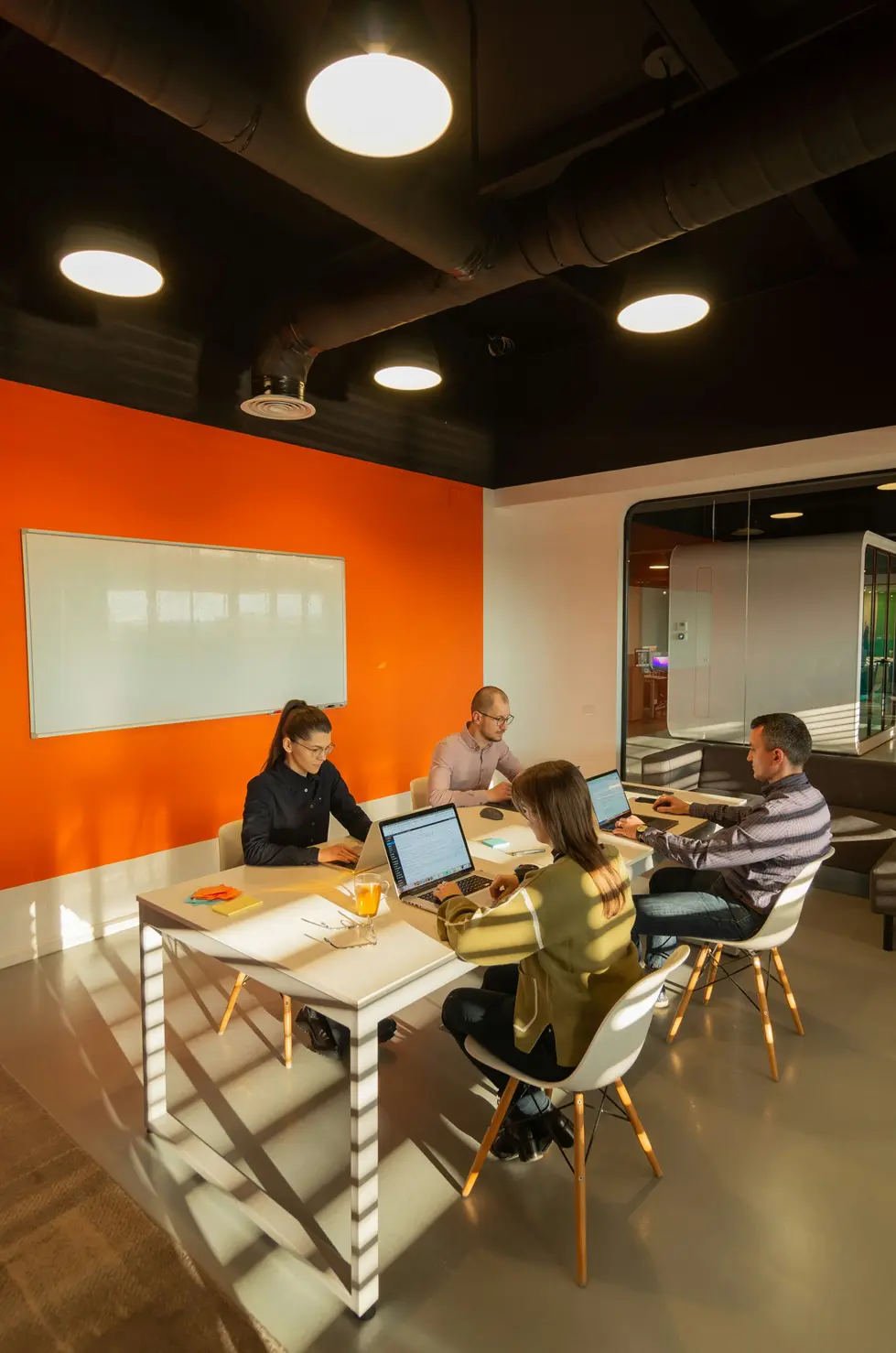 Four colleagues sit around a white table in a modern office, working on laptops near an orange accent wall and whiteboard.