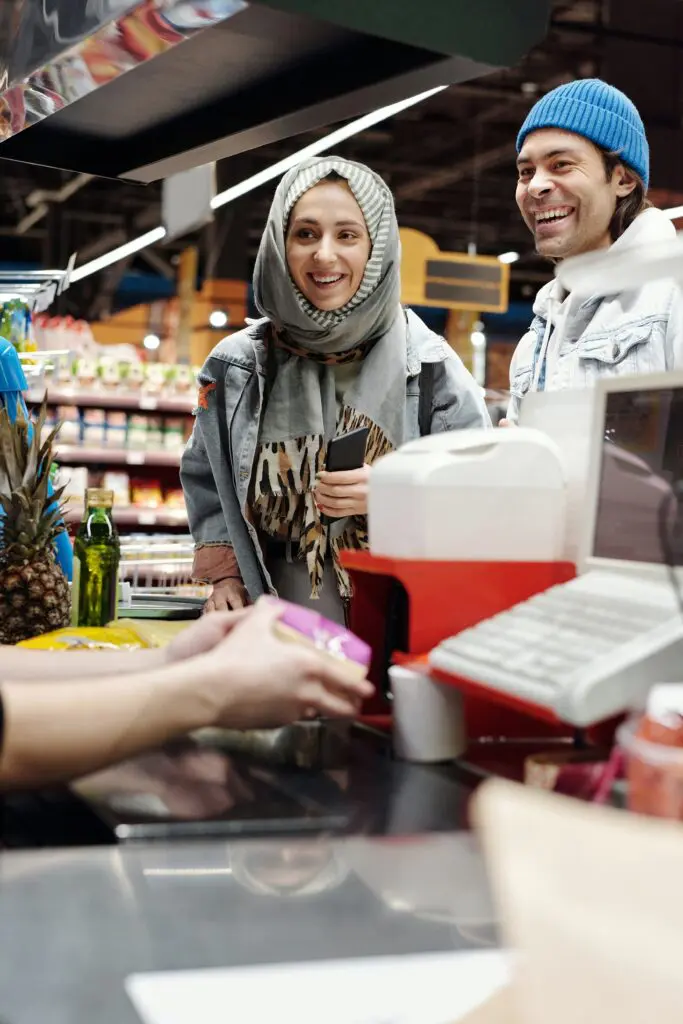 Smiling woman wearing a hijab and denim jacket at a checkout with a man in a blue knit cap beside her in a grocery store.