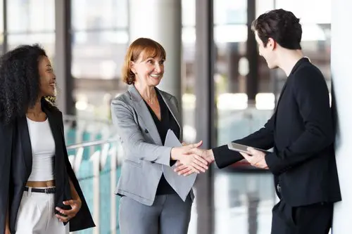 Three professionals in a modern office shake hands: a smiling woman in a gray suit (center) greets a man with a clipboard, with a woman in white nearby.