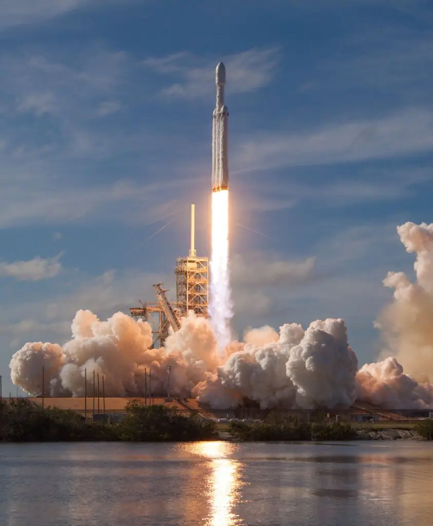 Rocket lifting off from launch pad, white exhaust plume and bright flame against blue sky. Mostly centered image with smoke billowing at base.
